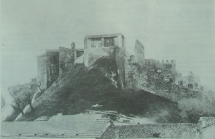 Portico delle campane comunali sul posto della torre crollata. A destra e sinistra le rovine dell'antica Rocca. In basso i tetti delle scuole.